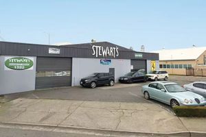 Stewart's auto repair shop with several cars parked outside. Gray building with a large sign, blue sky in background.